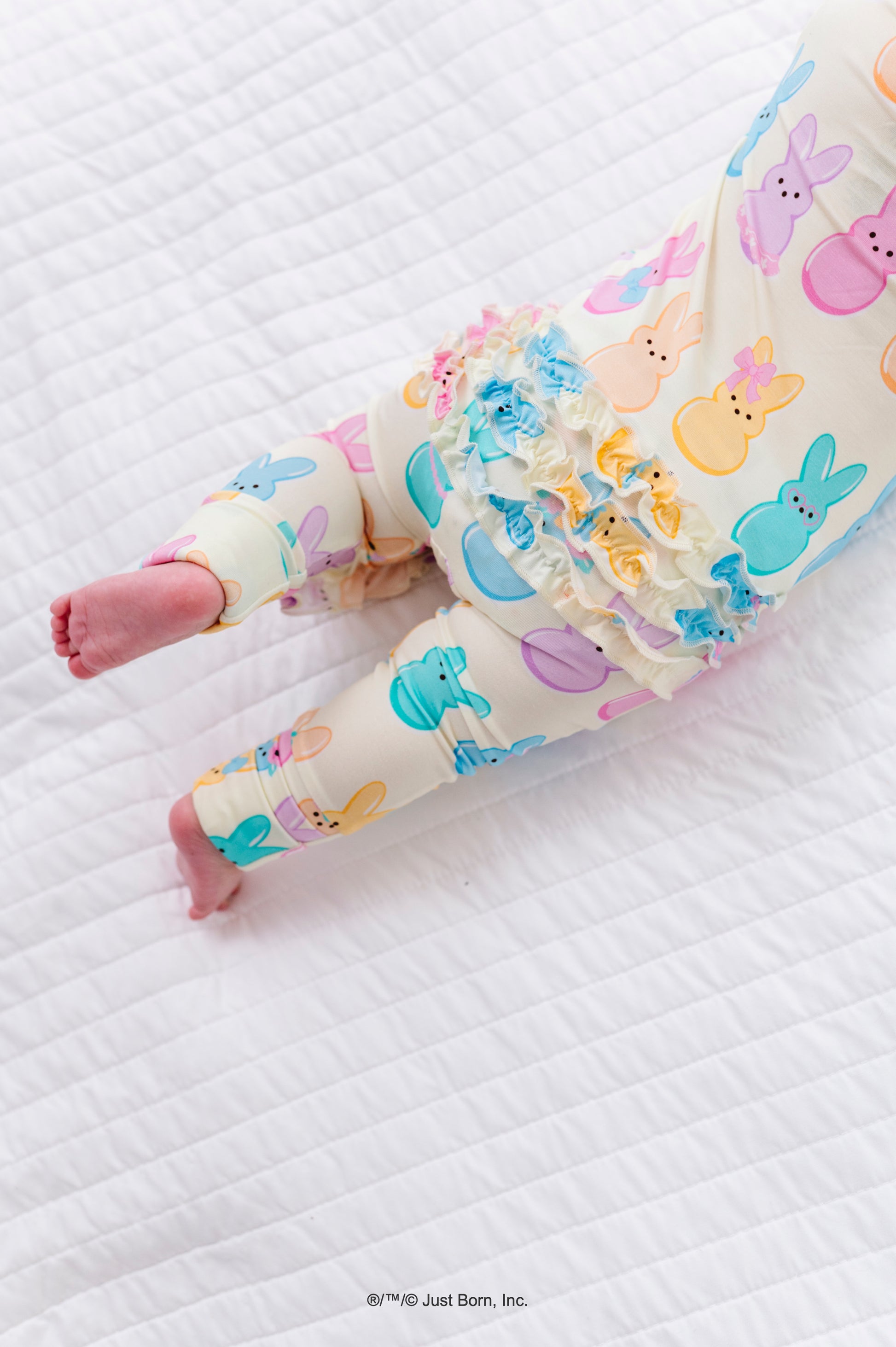 Baby legs wearing colorful romper with PEEPS® design on a white quilted surface.