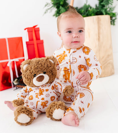 A baby wearing a white and orange holiday-themed onesie sits on a white floor next to a brown teddy bear, with red gift boxes and a green wreath in the background.