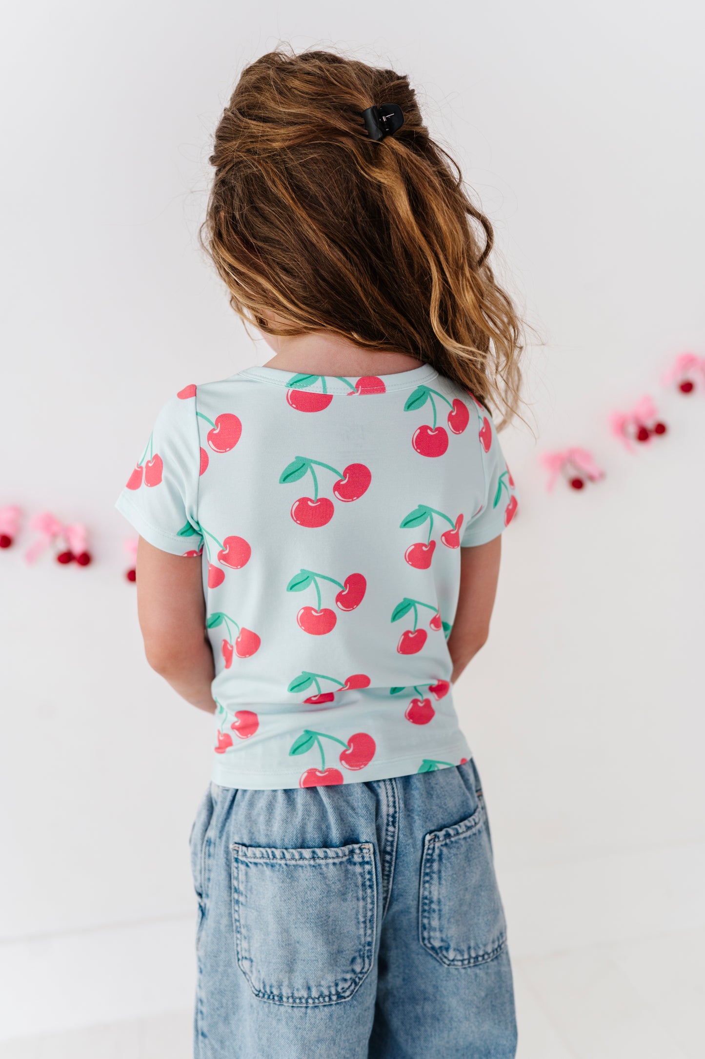 A child viewed from behind wearing a light blue short-sleeve t-shirt with a red cherry print and light blue denim jeans, standing against a white wall with pink garland.