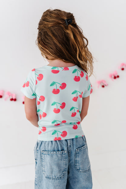 A child viewed from behind wearing a light blue short-sleeve t-shirt with a red cherry print and light blue denim jeans, standing against a white wall with pink garland.