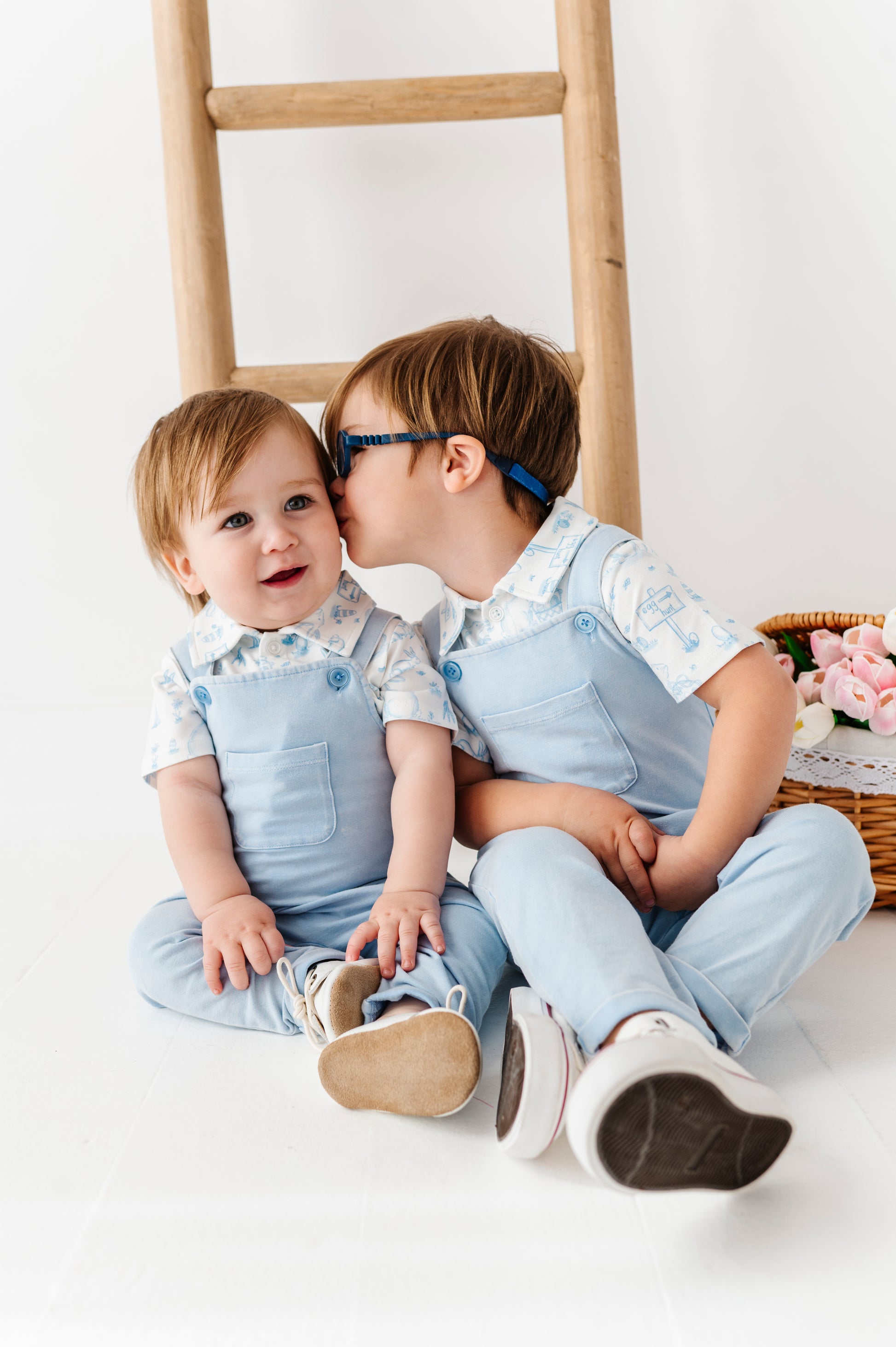 Two children in matching outfits sitting on a white floor with a wooden ladder in the background.