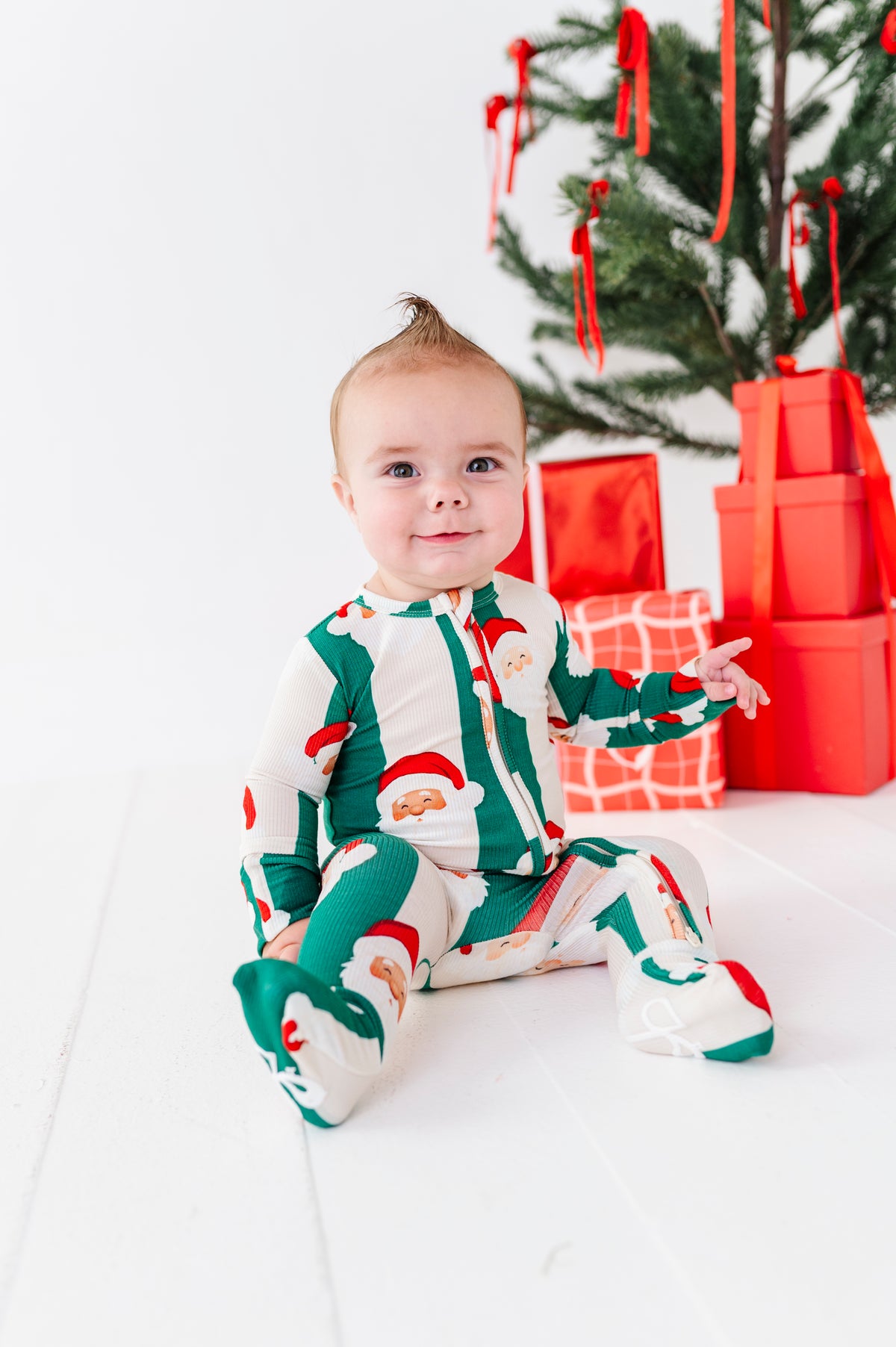 A baby sitting on a white floor in front of a Christmas tree and wrapped presents, wearing a green and white Santa Claus print footed bamboo pajama set.