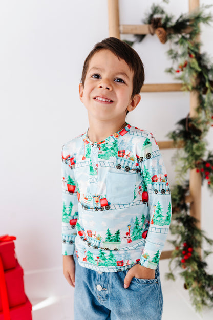 A young boy smiling and looking up, wearing a light blue long-sleeve bamboo henley shirt with a Christmas train and tree pattern, paired with blue jeans. The background is a bright white wall decorated with a wooden ladder and holiday greenery.