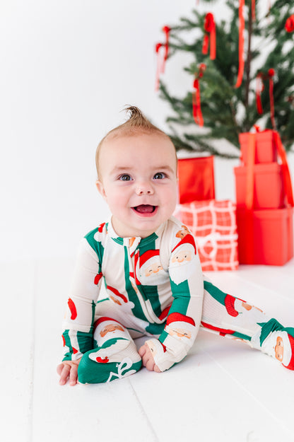 A baby wearing a festive Christmas-themed long-sleeve footed bamboo onesie sits smiling on a white floor in front of a decorated Christmas tree and wrapped gifts.