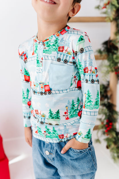 A child wearing a long-sleeve button-front bamboo shirt with a colorful Christmas train and tree print, paired with blue jeans, shown in a holiday-themed indoor setting.