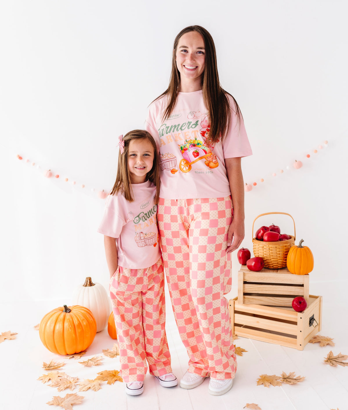 Two girls modeling matching pink pajamas in a styled studio setup with pumpkins, fall leaves, and a wooden crate. The longer girl wears a pink graphic T-shirt and pink-and-white checkered pants, while the shorter girl wears a matching pink T-shirt and pink-and-white checkered pajama pants.