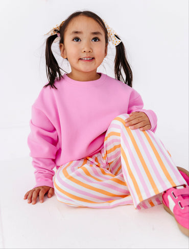 A young girl with dark hair in pigtails sits on a white floor wearing a pink sweatshirt and pink, orange, and white striped pants.