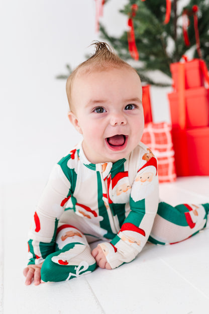 A baby wearing a festive Christmas Santa Claus print footed bamboo sleeper sits smiling on a white floor in front of a decorated Christmas tree and wrapped red gifts.