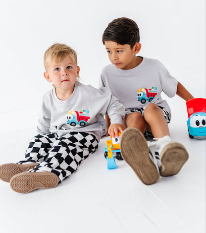 Two young boys sitting on a white studio floor wearing matching light gray long-sleeve t-shirts with a cartoon construction truck graphic on the chest. The boy on the left is wearing black and white checkered pajama pants, and the boy on the right is wearing dark shorts.