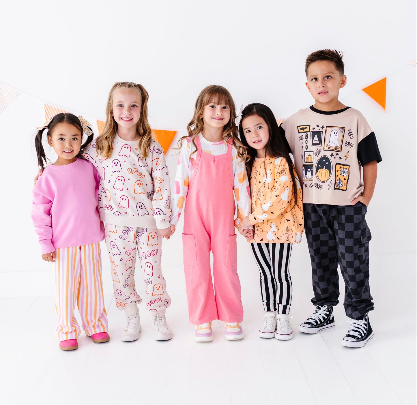 Five children standing together in a studio wearing matching pajama and casual outfits. The children are dressed in coordinated sets including pink, orange, white, and black patterns, with a white background and small orange triangle decorations behind them.