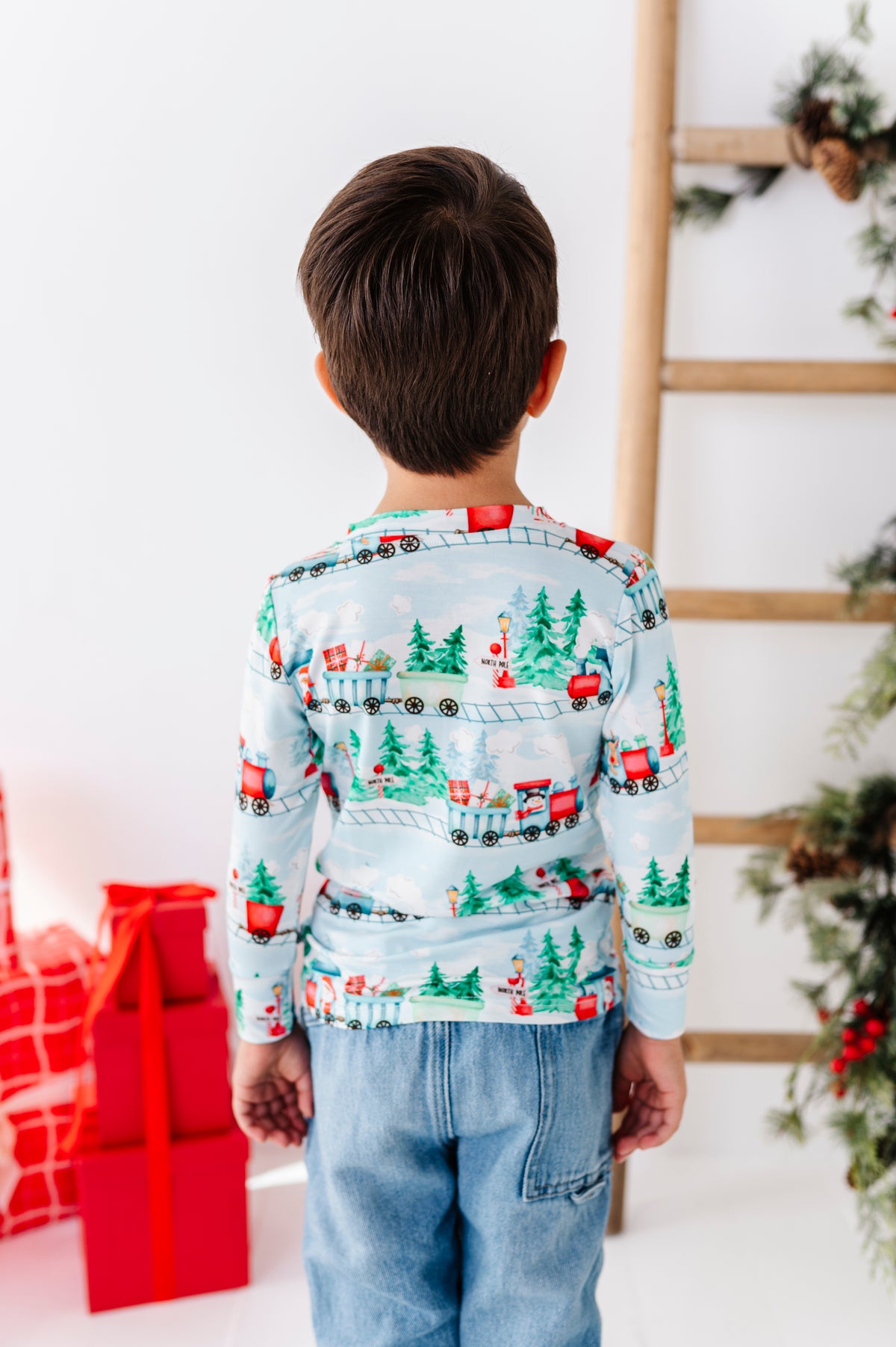A child viewed from behind wearing a light blue long-sleeve bamboo henley shirt with a Christmas train and tree print, paired with light blue jeans in a holiday-themed room.