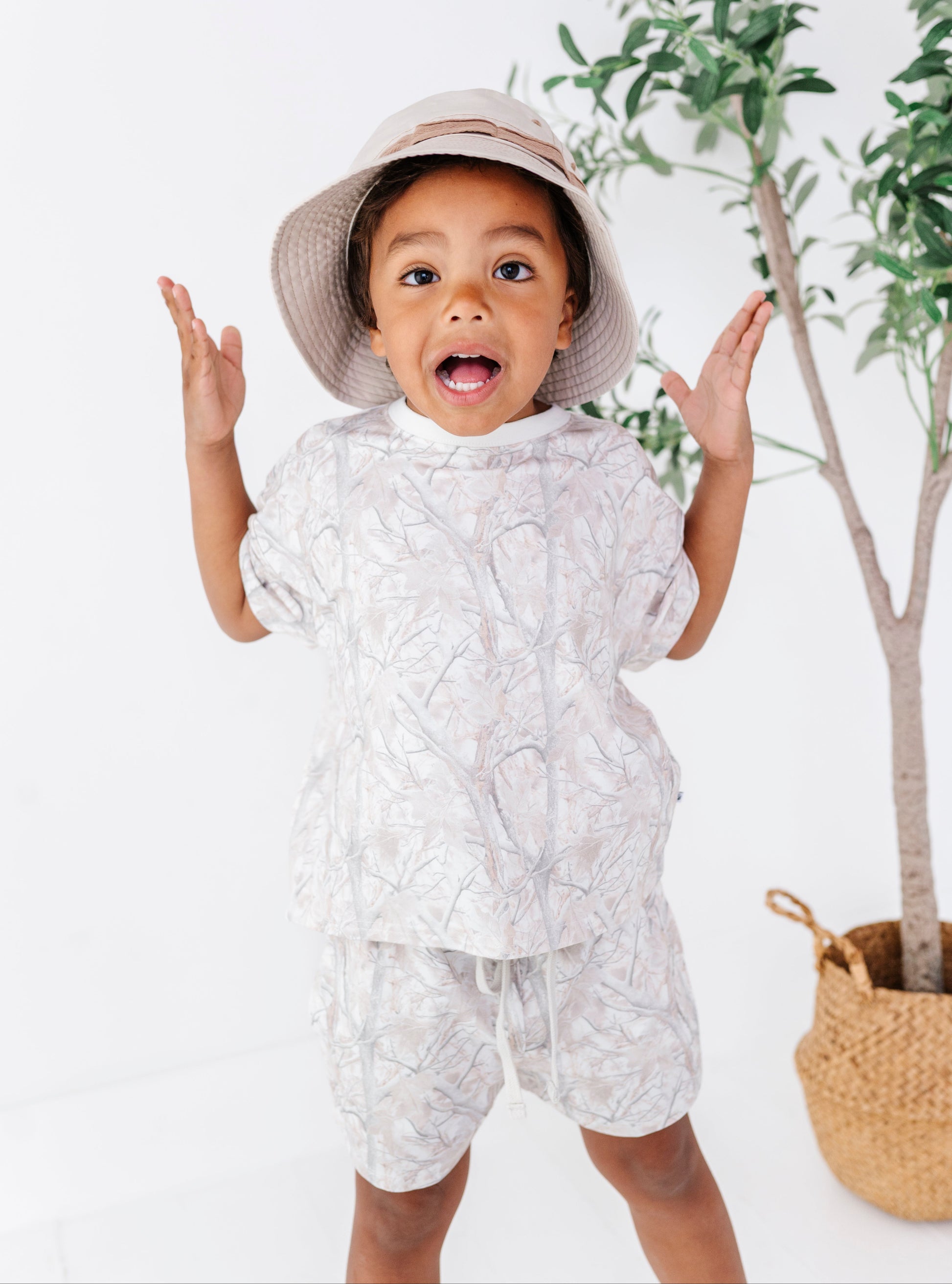 Child wearing a tree camo patterned outfit with a hat, standing in front of a plant on a white background