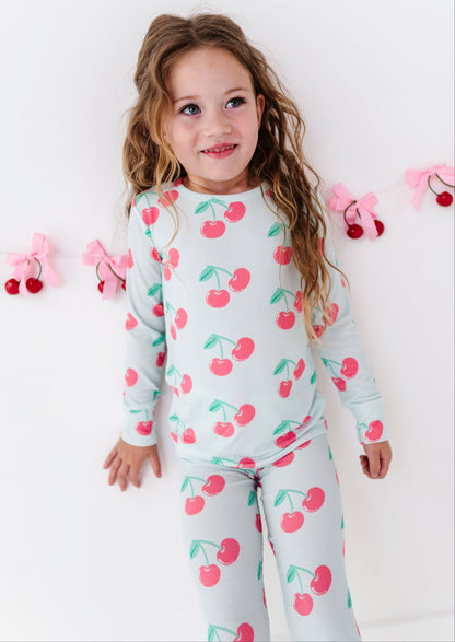 A young girl with long wavy light brown hair is wearing a light gray long-sleeve pajama set with a pink and green cherry print, standing against a white wall with pink bow and cherry decorations.