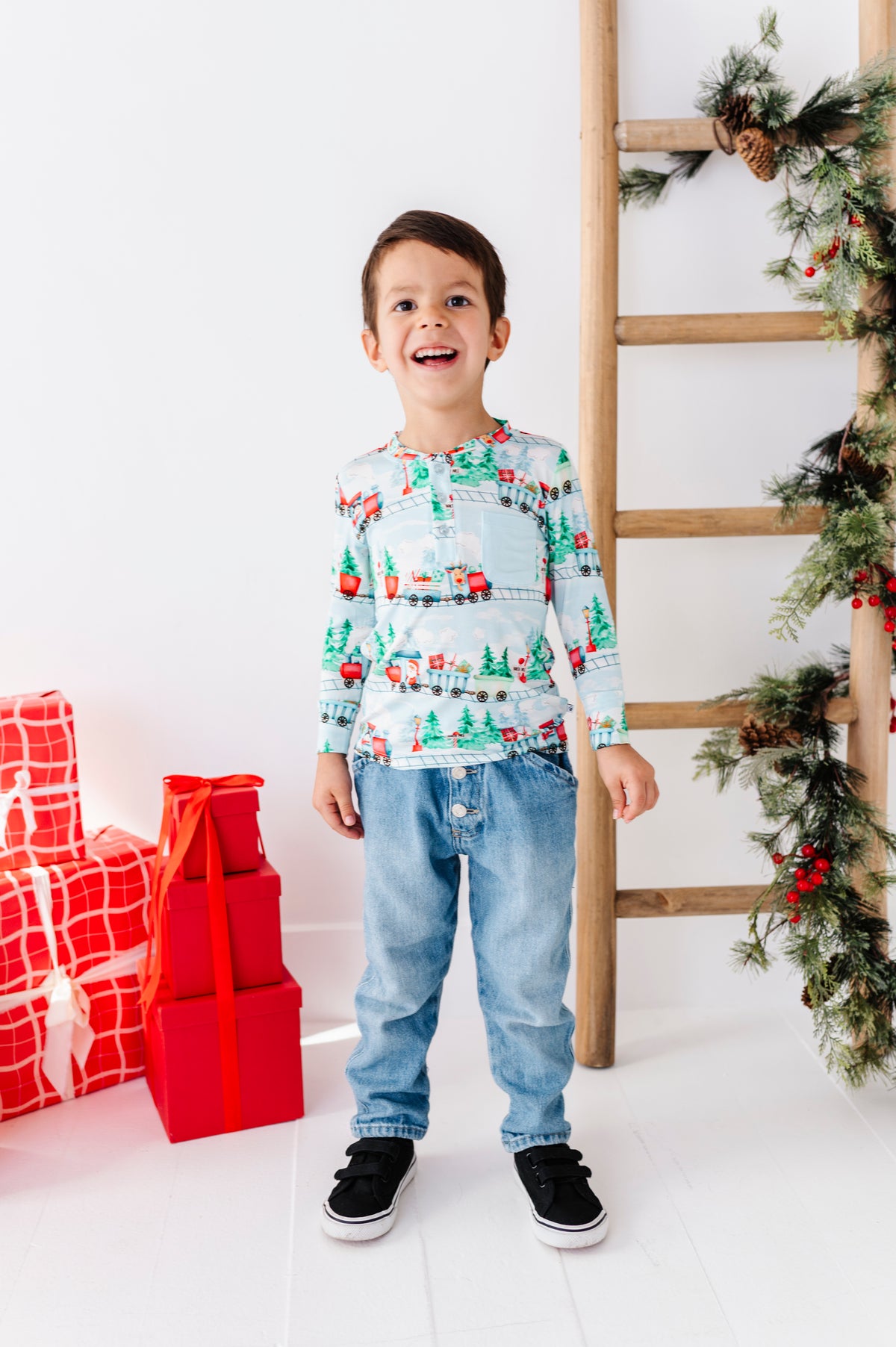 A young boy stands smiling in a holiday-themed room, wearing a long-sleeve crewneck shirt with a Christmas train pattern, blue jeans, and black sneakers. To his left are wrapped Christmas presents, and to his right is a wooden ladder decorated with pine garlands.