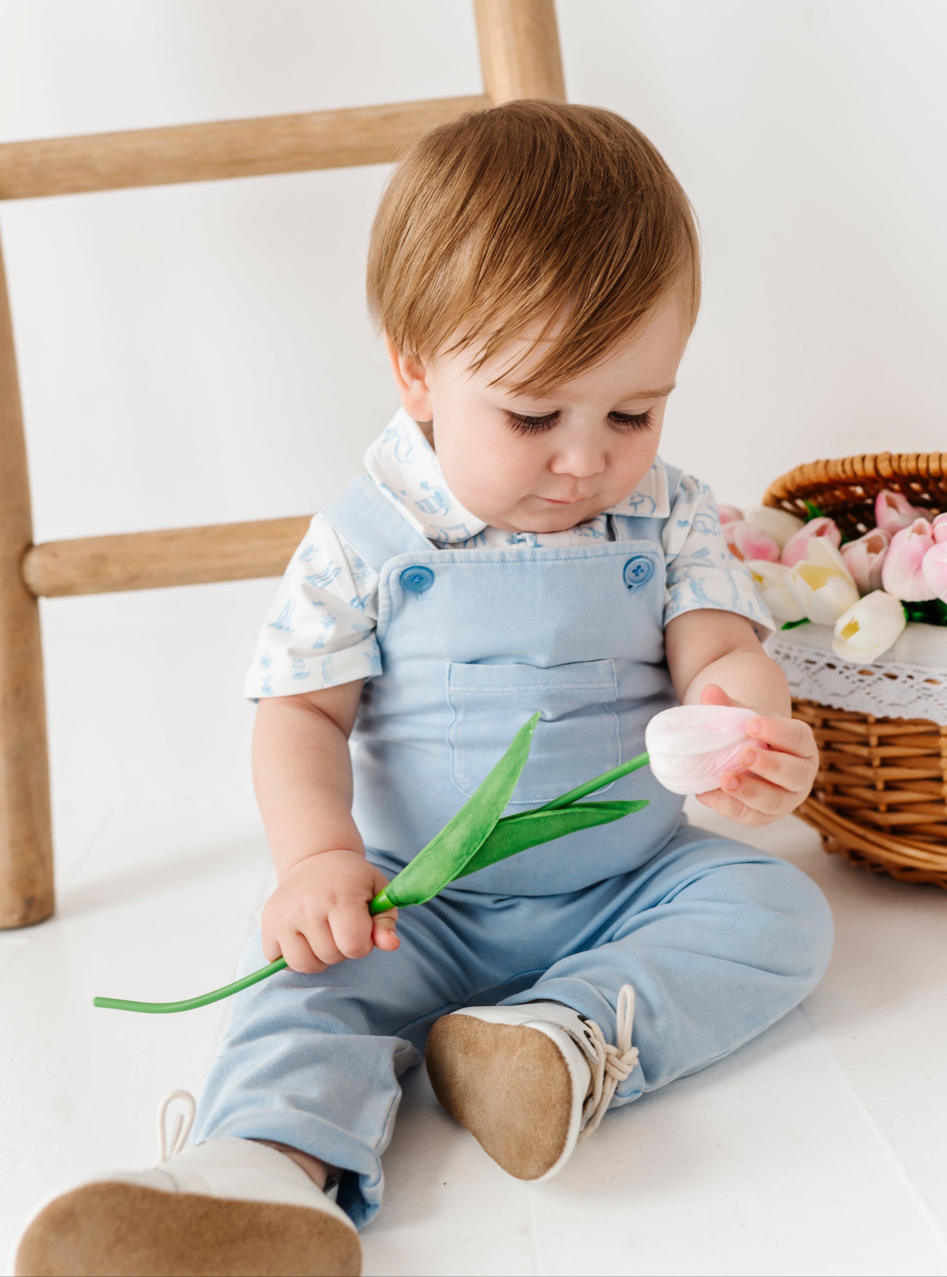 Child in denim overalls holding a tulip and an egg on a white floor with a wooden chair and basket of flowers in the background.