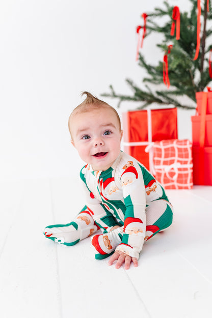 A baby sitting on a white floor in front of a Christmas tree and wrapped gifts, wearing a festive Santa Claus print one-piece bamboo pajama. The pajama features a cream base with green and red holiday patterns.