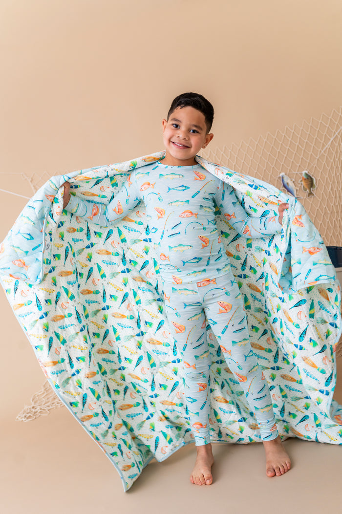 Child wearing a colorful fish-patterned bamboo pajama set with a matching blanket draped over them on a beige background.