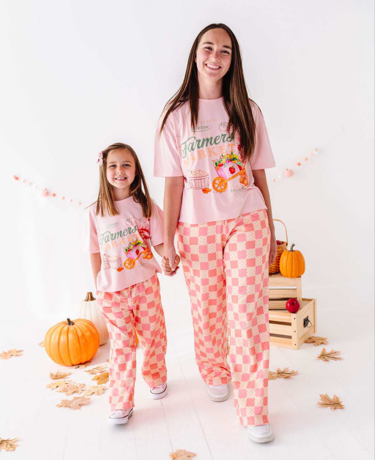 A woman and a young girl wearing matching light pink graphic pajamas with peach-and-white checkered pants, standing in a bright studio with pumpkins, fall leaves, and a wooden crate.