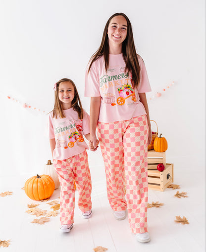 A woman and a young girl wearing matching light pink graphic pajamas with peach-and-white checkered pants, standing in a bright studio with pumpkins, fall leaves, and a wooden crate.