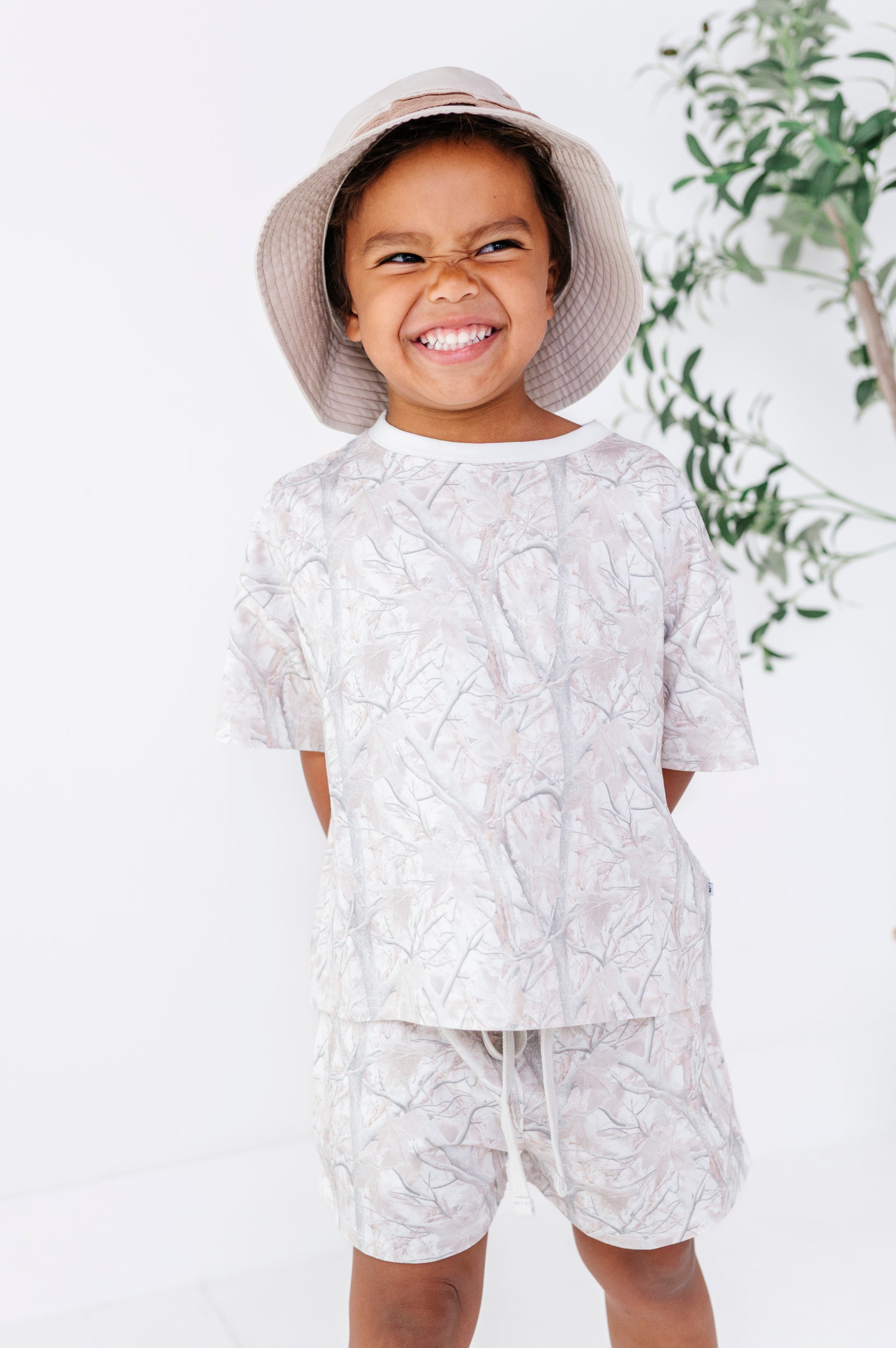 Child wearing a camo patterned outfit with a hat, standing against a white background with greenery.
