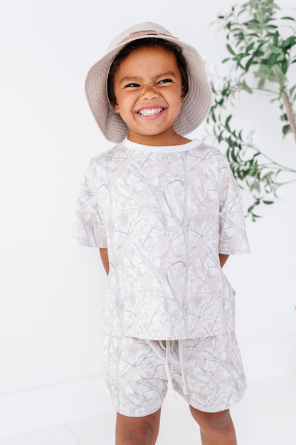 Child wearing a camo patterned outfit with a hat, standing against a white background with greenery.
