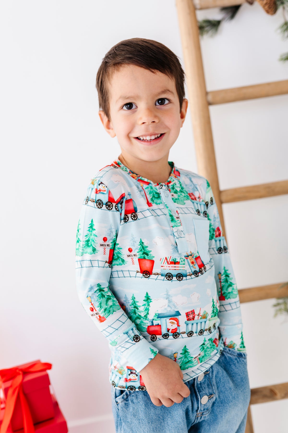 A child wearing a long-sleeve bamboo henley shirt with a festive Christmas train pattern, paired with blue jeans. The shirt features a light blue background with red, green, and white holiday illustrations of trains, pine trees, and snowflakes.