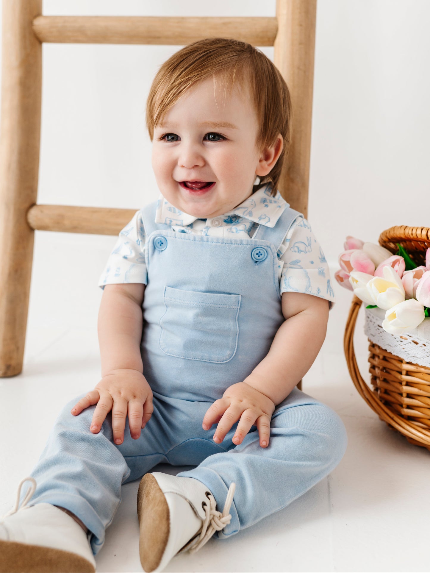 Baby wearing light blue overalls sitting on a white floor with a wooden ladder and basket of flowers in the background.