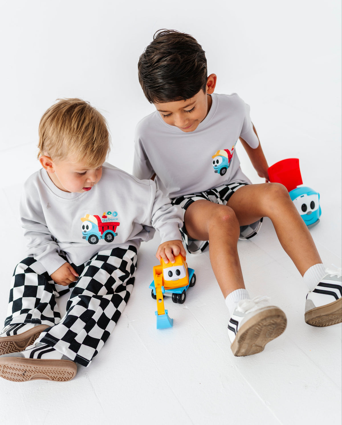 Two young children sitting on a white floor, wearing matching light gray long-sleeve shirts with a colorful truck graphic on the chest and black-and-white checkered pajama pants. One child is holding a small toy excavator.