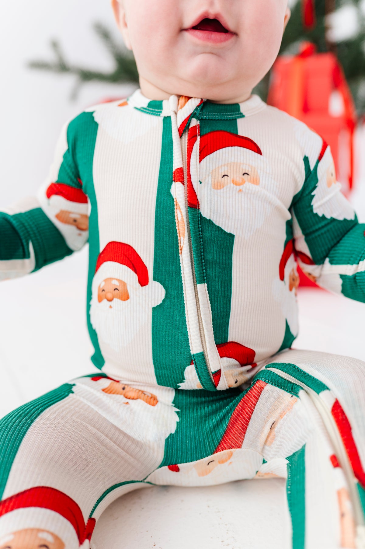 A baby wearing a festive Christmas-themed zip-up bamboo sleeper with a Santa Claus pattern, sitting against a soft white background with blurred holiday decorations.