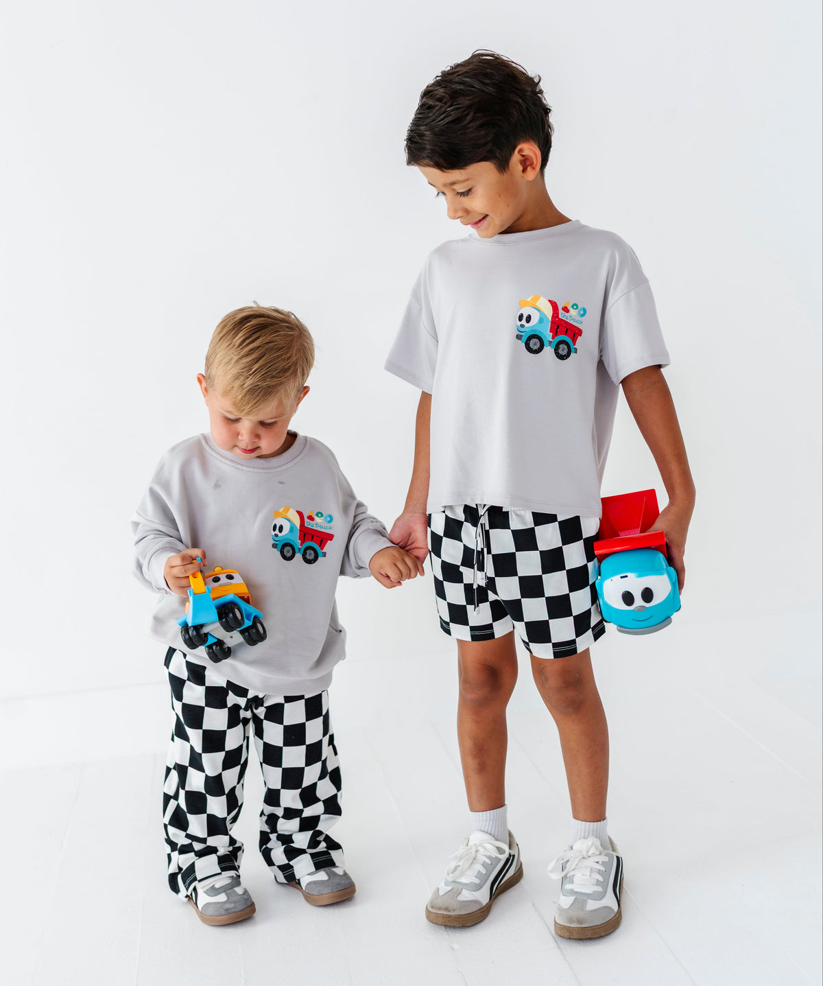 Two children modeling matching light gray crewneck t-shirts and checkered pants, standing in a bright white studio. The children are holding colorful toy trucks.