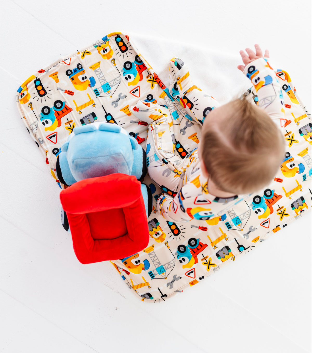 A baby lies on a colorful printed blanket with toy vehicles, alongside a red plush toy and a blue cup, on a white wooden floor.