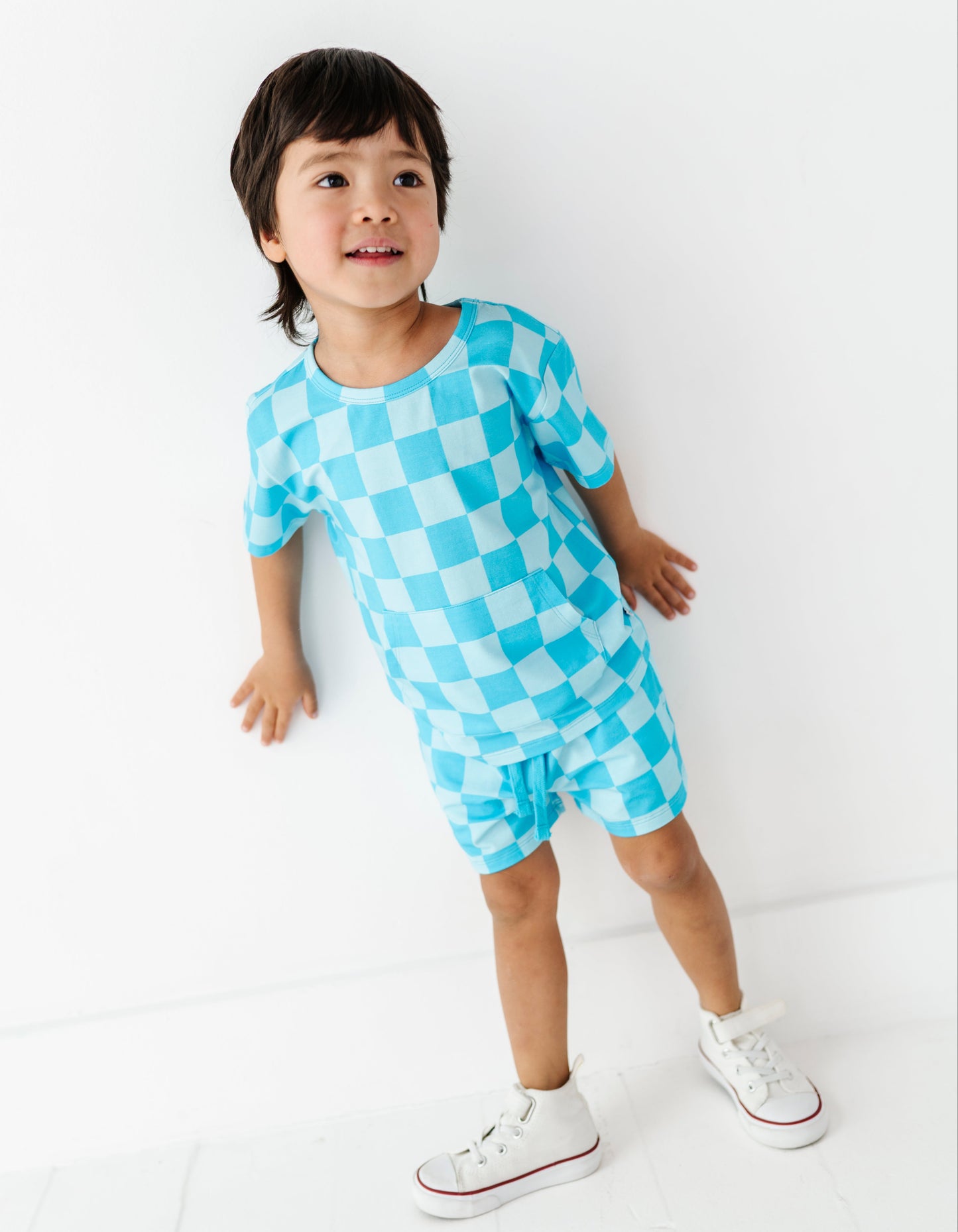 A young child wearing a light blue and white checkered short-sleeve romper and white sneakers, standing against a clean white wall.