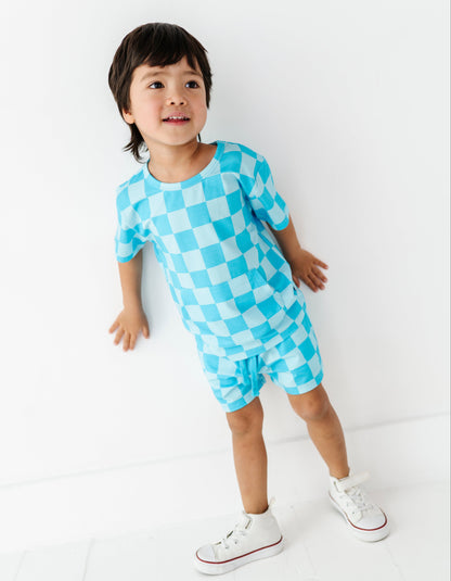 A young child wearing a light blue and white checkered short-sleeve romper and white sneakers, standing against a clean white wall.