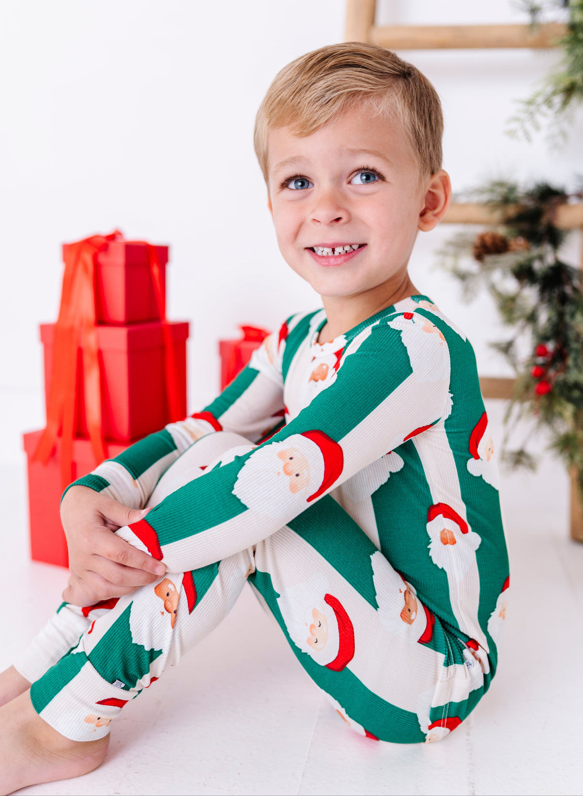 A young blonde boy wearing green-and-beige striped bamboo pajamas with Santa faces all over them. The super soft bamboo pajamas are ribbed and made to fit snugly for safety standards. He's sitting in front of a stack of red wrapped Christmas gifts 