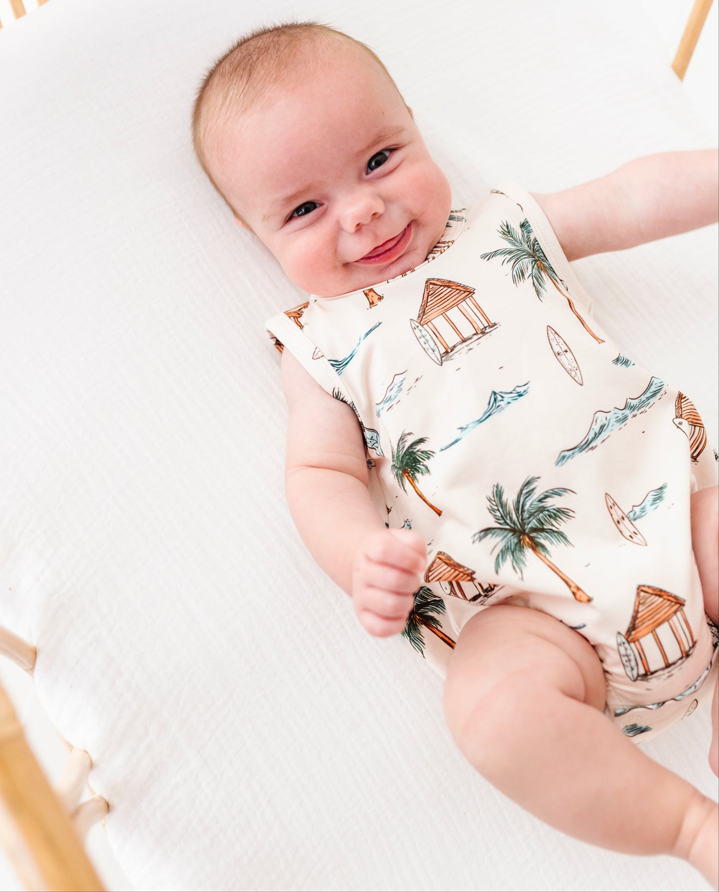 Baby lying in a crib wearing a sleeveless romper with beach-themed print.