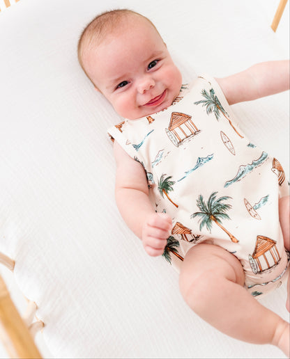 Baby lying in a crib wearing a sleeveless romper with beach-themed print.