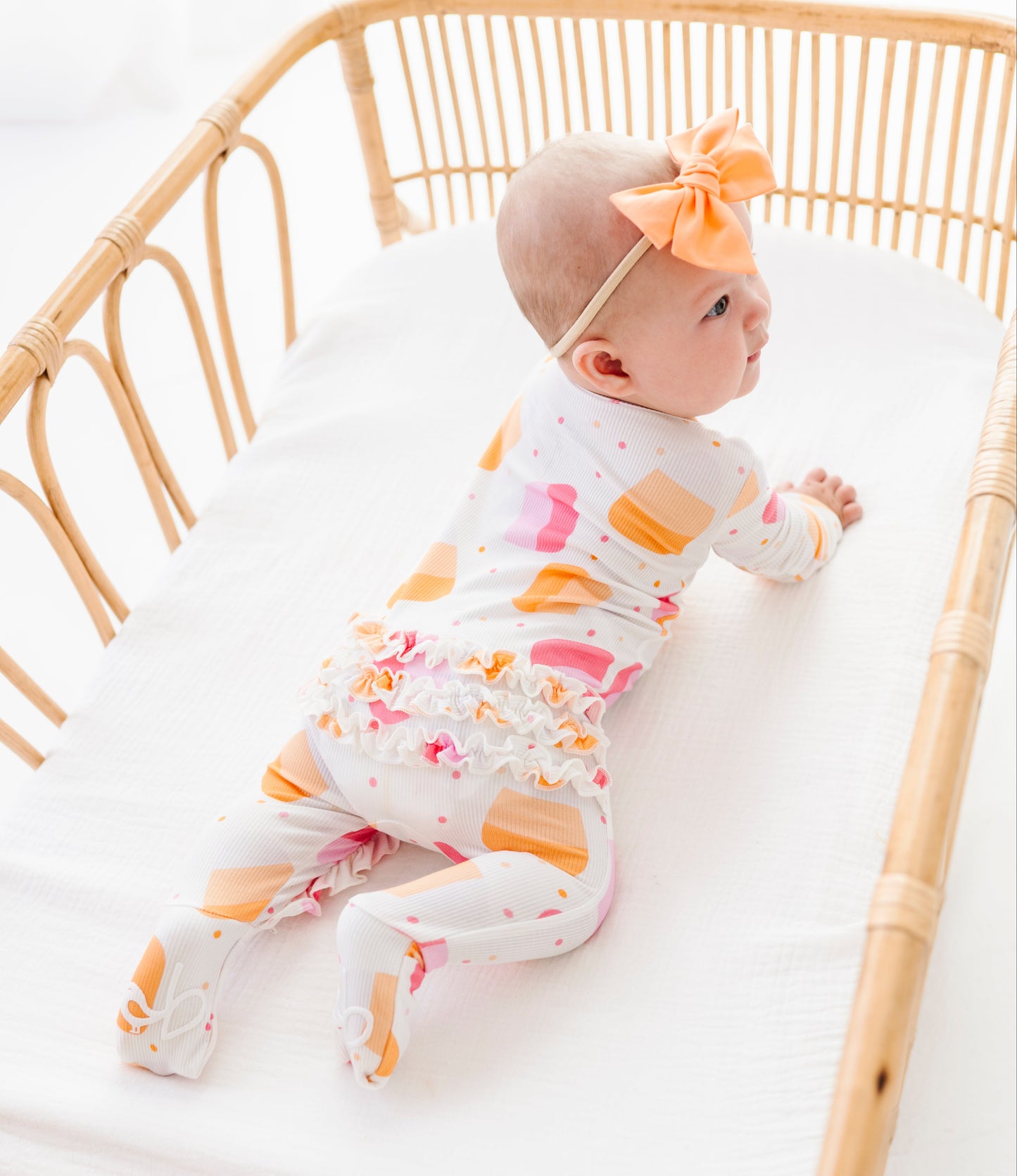 A baby lying on their stomach in a light wood rattan bassinet, wearing a white footed sleeper with a colorful candy print and a matching orange bow headband.