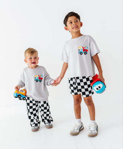 Two children modeling matching white t-shirts and black-and-white checkered pants/shorts in a bright studio setting. The larger child holds a blue toy, and both wear matching outfits with a small colorful truck graphic on the chest.