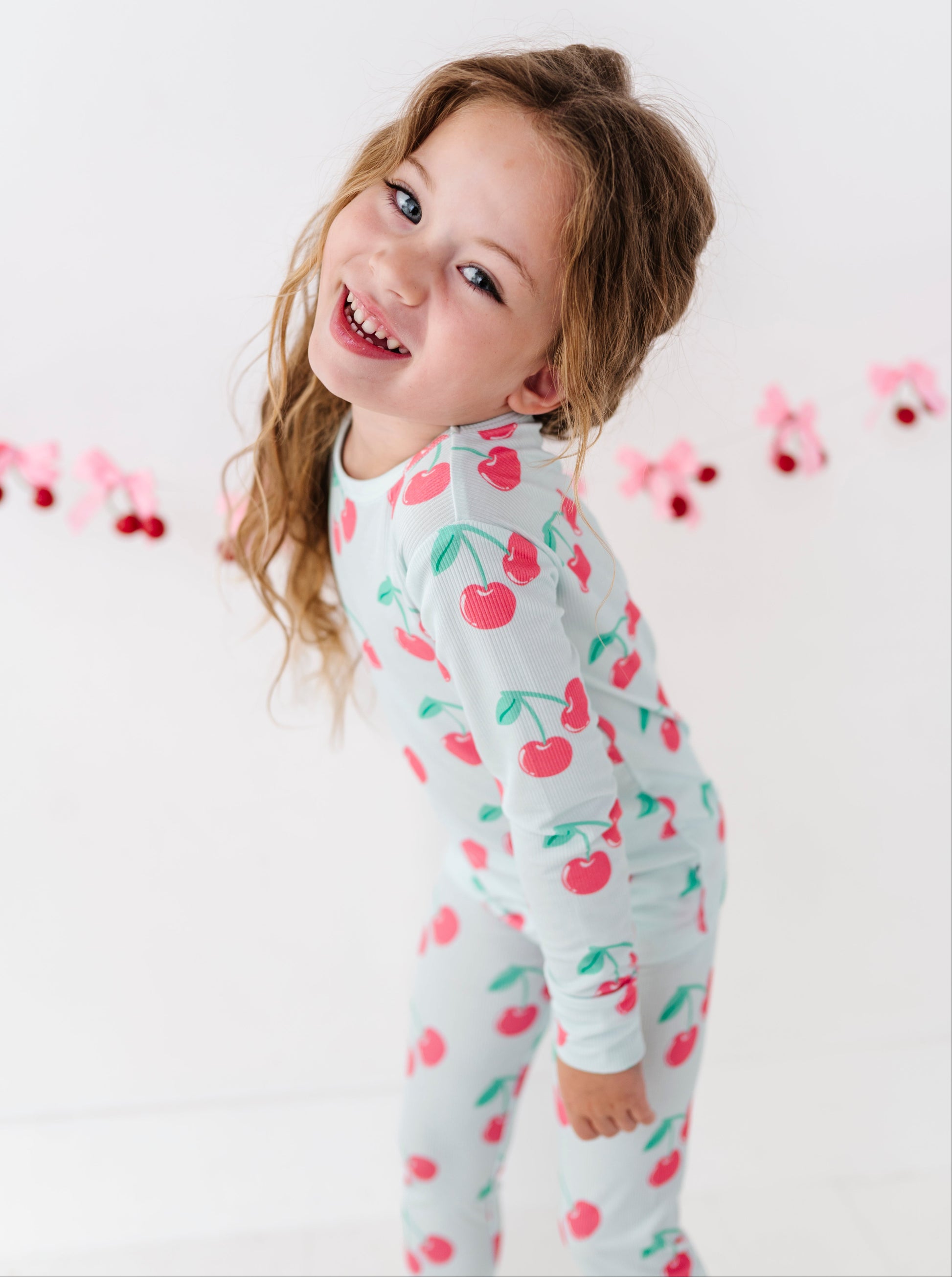 A young girl with light brown hair smiling and looking over her shoulder, wearing a white long-sleeve pajama set with a red cherry and green leaf pattern. She is standing against a white background with a pink and red cherry garland hanging in the distance.
