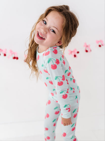 A young girl with light brown hair smiling and looking over her shoulder, wearing a white long-sleeve pajama set with a red cherry and green leaf pattern. She is standing against a white background with a pink and red cherry garland hanging in the distance.