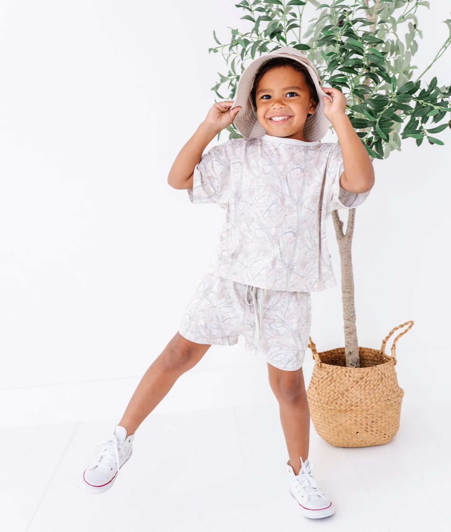 Child wearing a light-colored tree camo outfit with a plant in the background