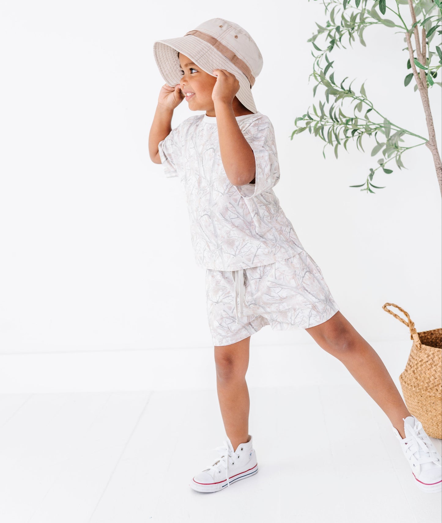 Child wearing a white outfit with a pattern, standing next to a plant on a white background