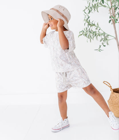 Child wearing a white outfit with a pattern, standing next to a plant on a white background