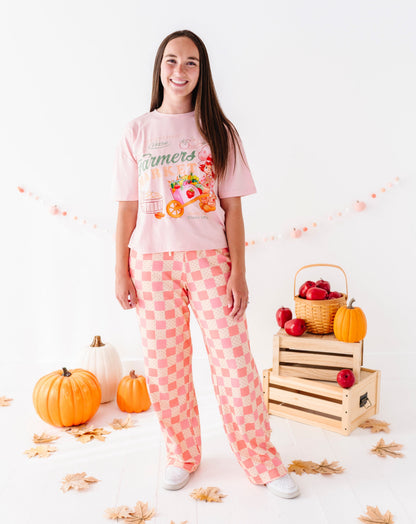 A young woman stands in a studio wearing a light pink graphic T-shirt and a matching pink-and-white checkered pajama set. The scene is styled with pumpkins, fall leaves, wooden crates, and a basket of red apples against a clean white background.