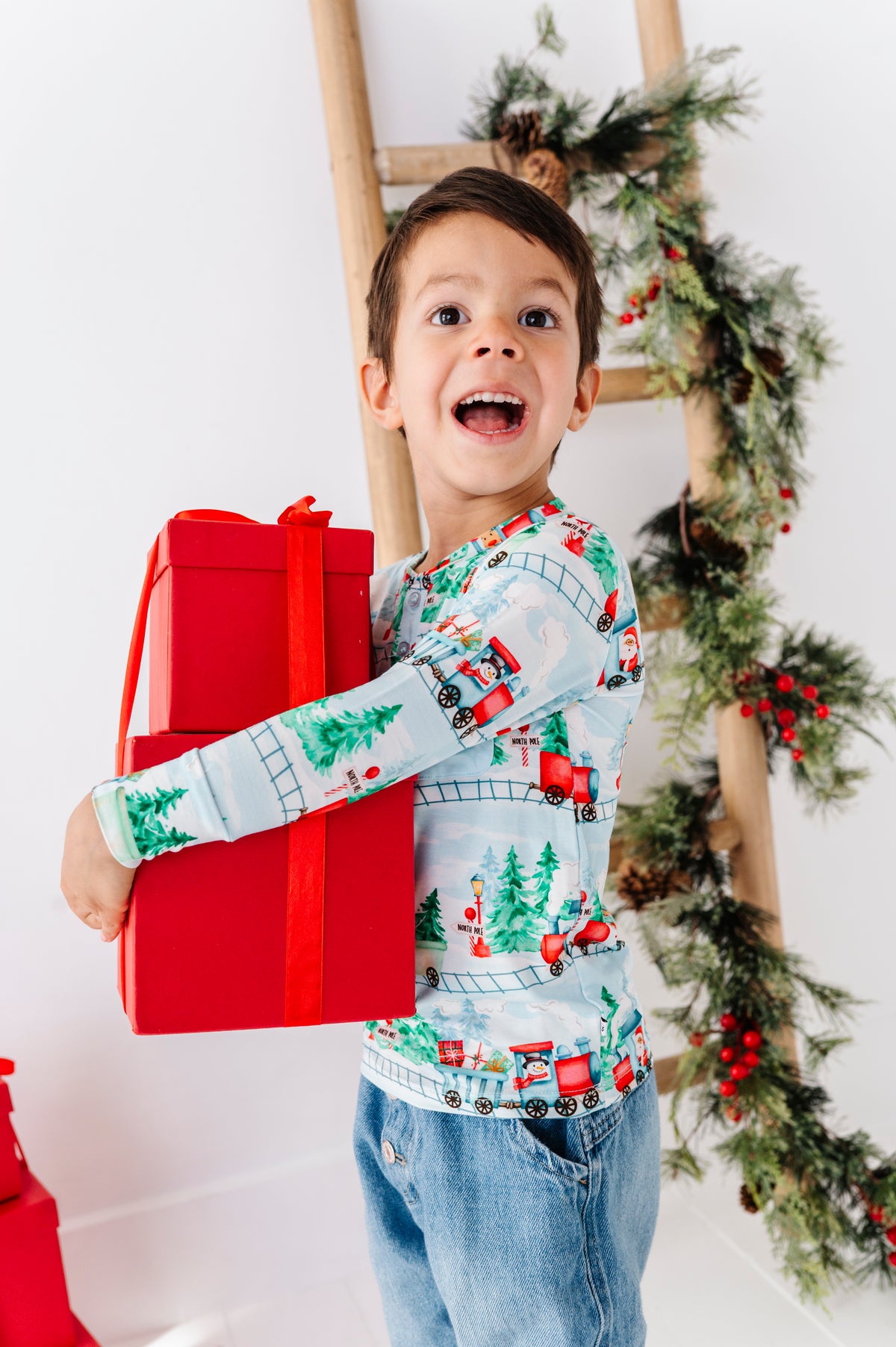 A child wearing a long-sleeve bamboo henley shirt with a festive Christmas train pattern, paired with blue jeans. The shirt features a light blue background with red, green, and white holiday illustrations of trains, pine trees, and snowflakes. He's holding a stack of red-wrapped presents and standing in front of a Christmas tree. 