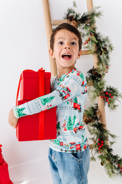 A child wearing a long-sleeve bamboo henley shirt with a festive Christmas train pattern, paired with blue jeans. The shirt features a light blue background with red, green, and white holiday illustrations of trains, pine trees, and snowflakes. He's holding a stack of red-wrapped presents and standing in front of a Christmas tree. 