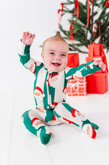 A baby sitting on a white floor in front of a Christmas tree and wrapped gifts, wearing a green and white Santa Claus print bamboo footed pajama romper.