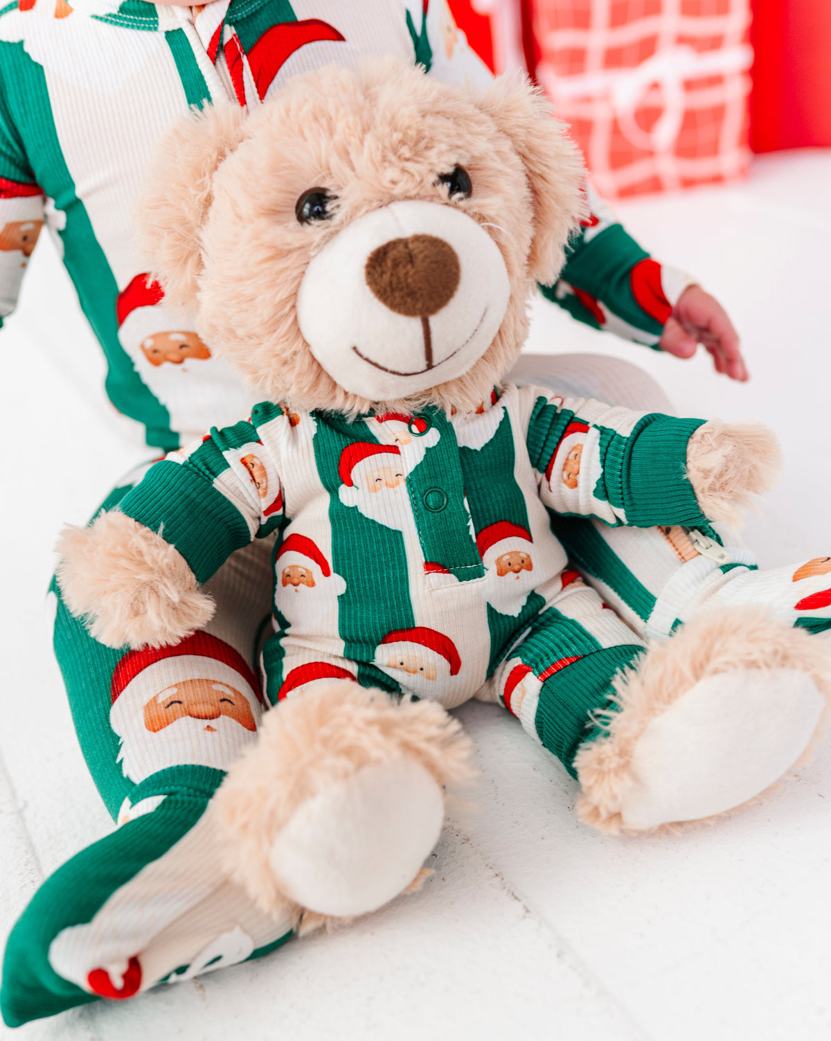 A plush teddy bear wearing festive green and white Christmas pajamas with Santa Claus patterns, posed on a white surface with a blurred person in matching pajamas in the background.