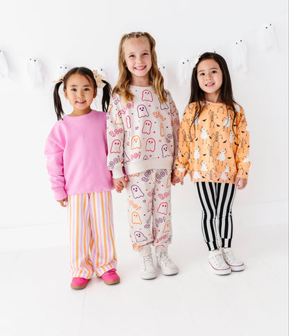Three young girls standing together in a bright white studio, wearing halloween clothing sets. The girl on the left wears a pink long-sleeve top with yellow and white striped pants.