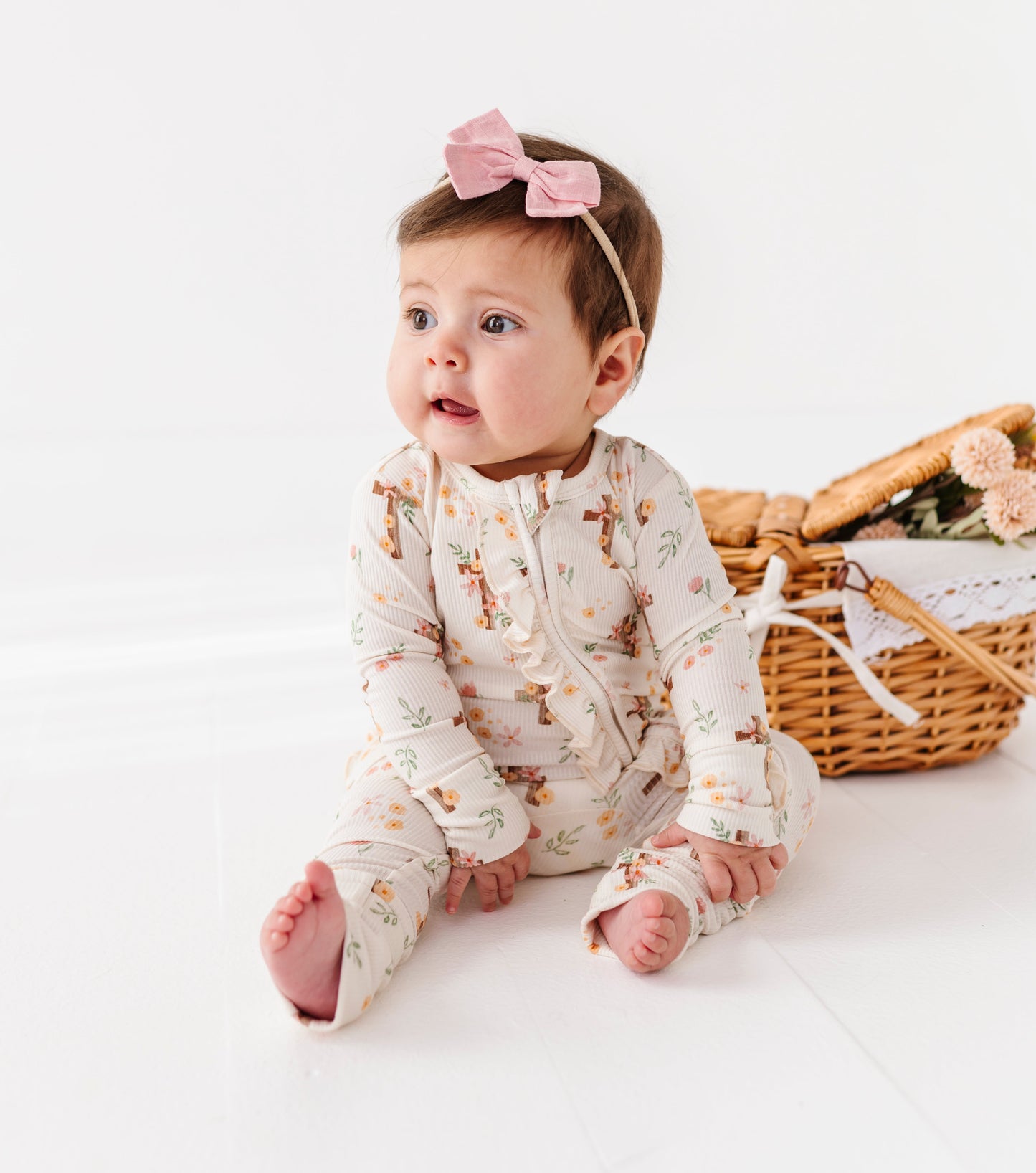 Baby wearing a floral bamboo onesie with a pink headband, sitting next to a wicker basket on a white background.